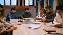 Group of people sitting around a wooden table all with books open in front of them and looking down at the books