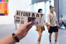 Hand wearing a watch holding out art/museum tickets with a large room with painting on the wall in the backgroun